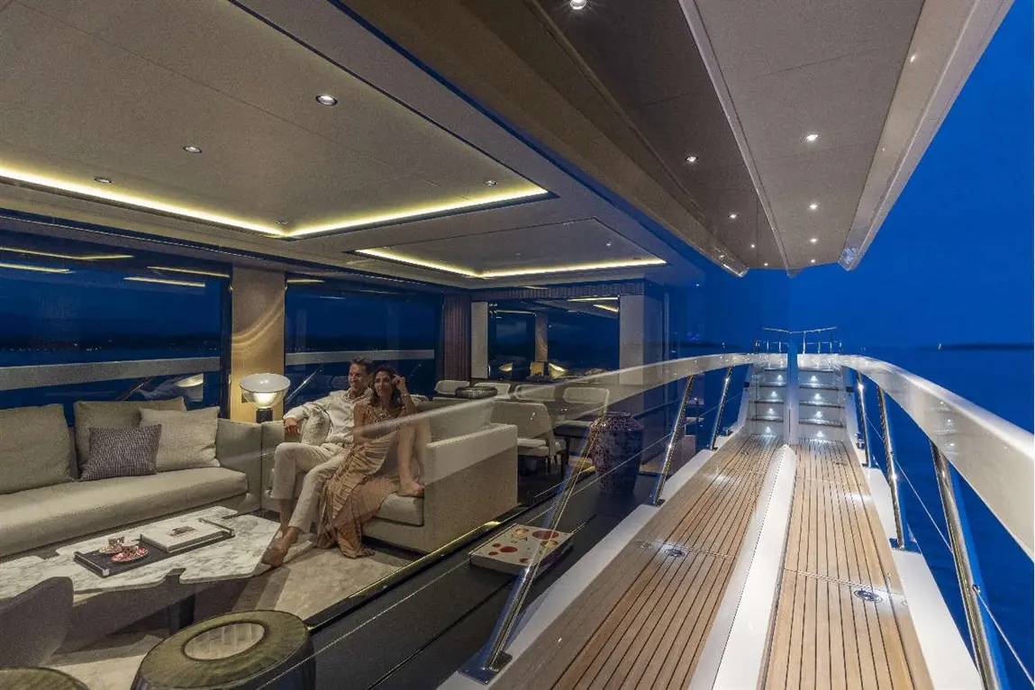 A couple relaxes on a luxurious yacht's deck at dusk, with elegant furnishings and calming ocean views beyond the glass.